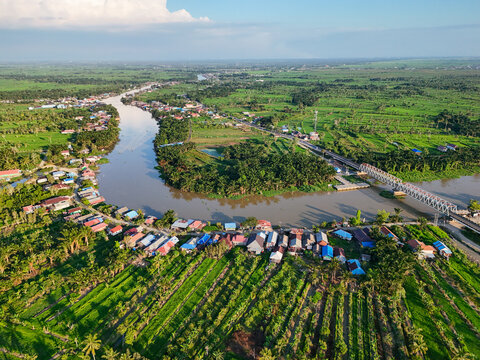 Aerial view of the river winding through the village, surrounded by lush green rice fields and a bridge crossing the water in Sungai Tabuk, South Kalimantan, Indonesia.