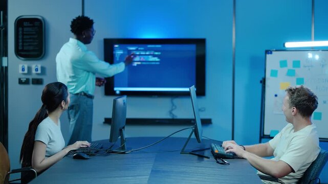 Diverse IT team of software developers coding and collaborating in a modern tech office. African American manager points at screen explaining project workflows to programmers working on computers.