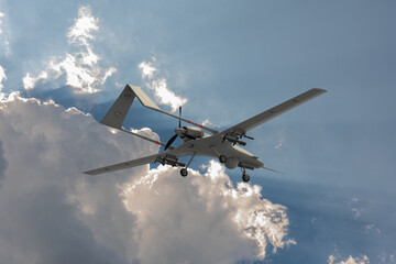 Obraz premium Military drone in flight with mounted munitions, optical equipment, propeller and extended landing gear against dramatic clouds.