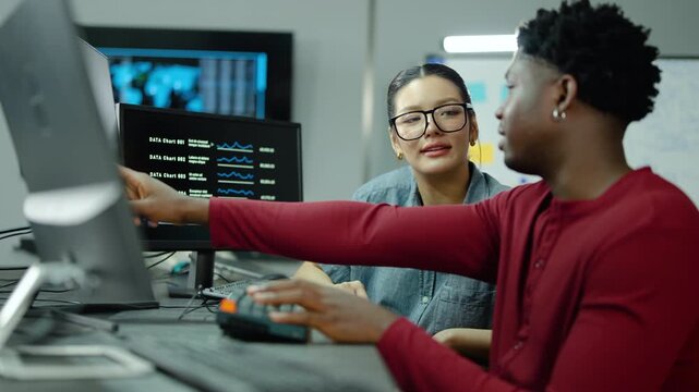 Two diverse data analysts reviewing financial charts on dual monitors in a high tech office. Smiling professionals collaborating on digital statistics and business growth strategy late at night.