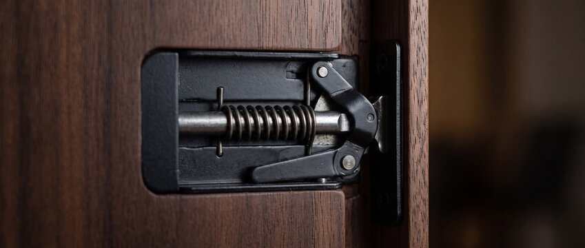 Close-up of metal spring latch hinge on wood door cabinet showing mechanism and hardware detail
