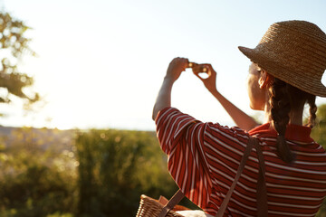 Obraz premium A young woman lifestyle tourist takes photos with a camera in a sunny field. She wears a hat and enjoys summer travel during golden hour. Concept for freedom, leisure and exploration.
