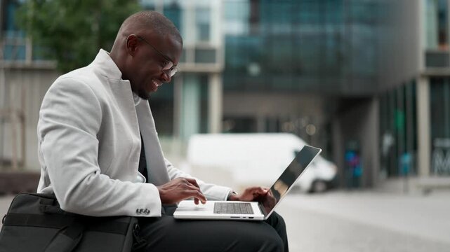 African american businessman, sitting, using laptop, analyzing data, outdoor near office building. Black business man smiling, works as it consultant, focused on daily tasks, collaborating. Copy space