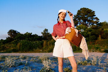Obraz premium Young woman lifestyle posing with a wicker basket and binoculars outdoors. The lady in vintage clothes enjoys a sunny day at the seaside. Concept for recreation and travel.