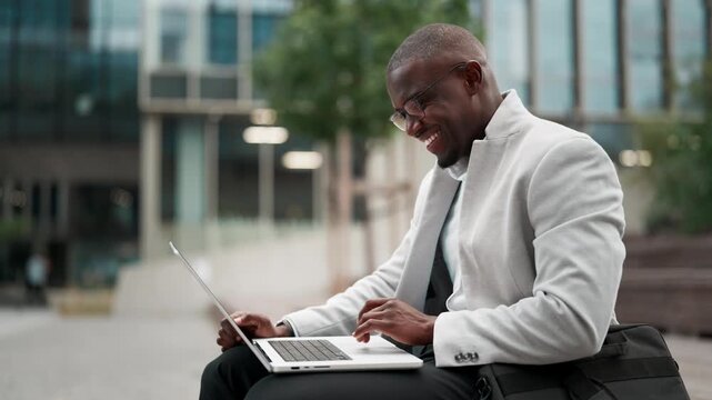 Black businessman, sitting, using laptop, reviewing budget, outdoor near office building. African american business man smiling, works as business analyst, prepared for client meeting, collaborating.