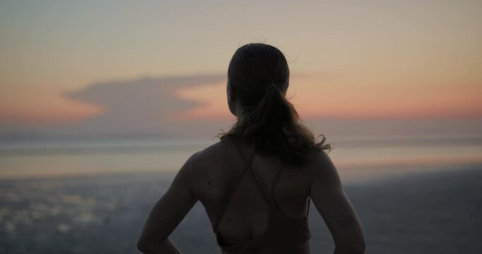 Slim woman with hands on hips looks at sunset ocean from beach backside view. Young female rests after yoga training on picturesque sea coast. Wellbeing