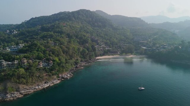 Crystal clear blue ocean water meets a sandy beach with green mountains in Phuket Thailand during a sunny day of tourism and relaxing travel