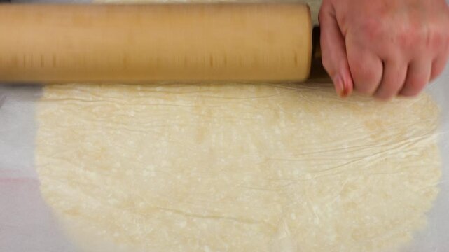 A female chef rolls out cottage cheese dough under a sheet of parchment paper using a wooden rolling pin, preventing the sticky dough from sticking and creating an even layer for home baking.