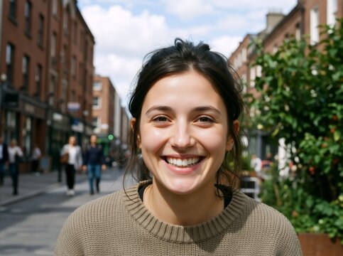 Portrait of a happy young brunette woman smiling on a city street. Cheerful female in a brown knit sweater outdoors. Urban lifestyle and travel concept