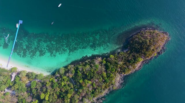 Bright turquoise waters surround a lush green island with sandy shores and a wooden pier in Phuket Thailand under clear sunny skies during the daytime