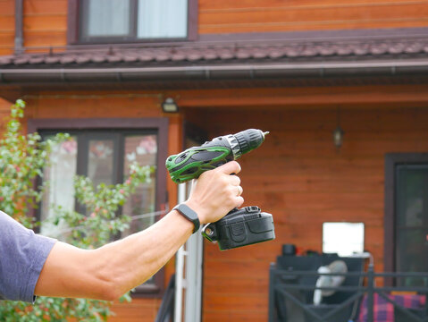 Arm and hand wearing a smartwatch holding a green cordless drill in front of a wooden cabin wall with a window.