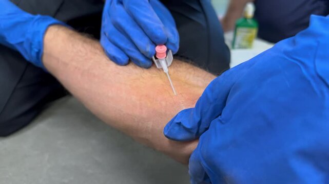 Gloved hands place an intravenous catheter into a forearm vein during paramedic skills training in a wartime setting. Faces not shown