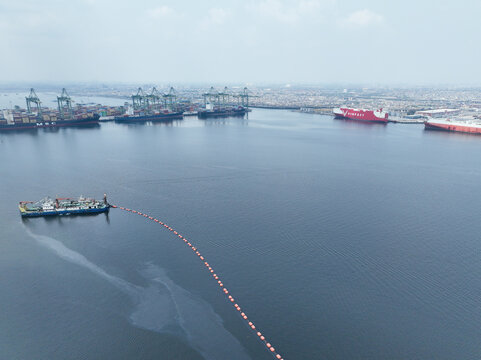 Aerial view of the busy Kali Baru port in Cilincing, featuring container ships, industrial cranes, and a vessel deploying a floating barrier, Cilincing, Jakarta, Indonesia.