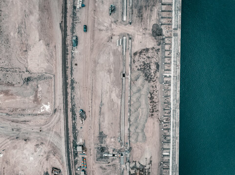 Aerial view of the Kali Baru port area featuring industrial storage piles, concrete infrastructure, and dirt ground along the waterfront in Cilincing, Jakarta, Indonesia.