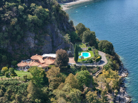 Aerial view of Capo Fornaci featuring a villa with a swimming pool nestled on a cliffside surrounded by lush trees and rocky terrain overlooking the water in Ispra, Lombardy, Italy.