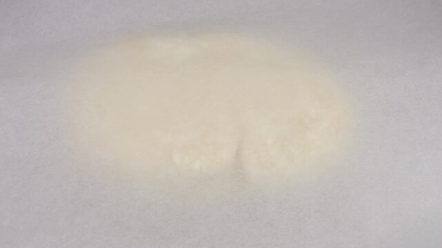 A lump of fresh cottage cheese dough with visible white flecks rests on a marked silicone mat, and a female chef rolls it out under parchment paper with a rolling pin to prevent sticking.