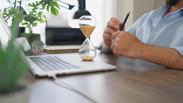 Businessman work on laptop with an hourglass and pen on office desk, planning important tasks and managing valuable time. Time management, productivity, business planning, workplace efficiency concept