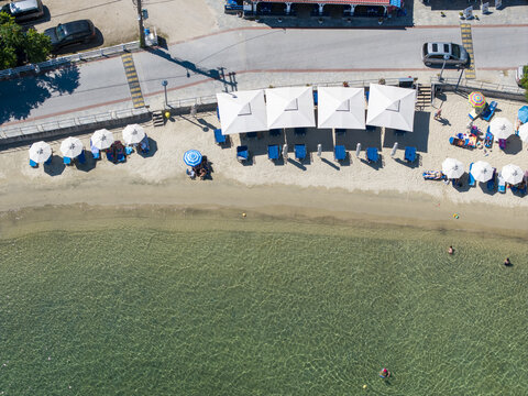 Aerial view of Sykias Beach with rows of white umbrellas and sun loungers on the sandy shore, bordered by clear turquoise water and a nearby road in Paralia Sikias, Greece.
