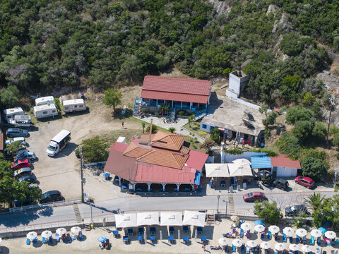 Aerial view of Sykias Beach featuring a restaurant with red tiled roofs, a parking area with cars and caravans, and beach umbrellas along the sandy shore in Paralia Sikias, Greece.