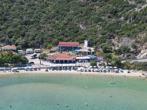 Aerial view of Sykias Beach with clear turquoise water, sun loungers, umbrellas, a waterfront restaurant, and lush green hills in the background in Paralia Sikias, Greece.