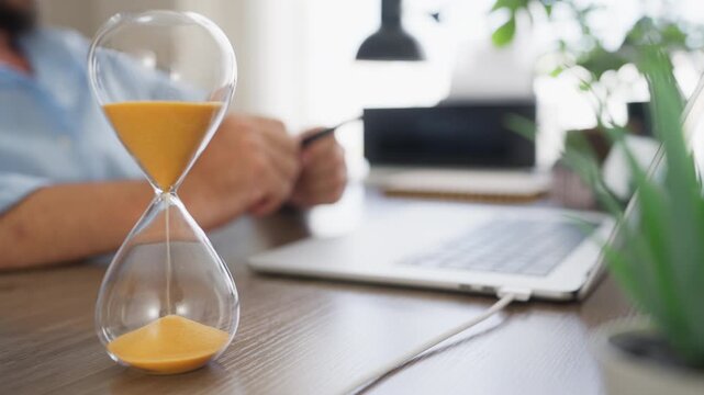Hourglass in sharp focus with a businessman working on a laptop in the background, symbolizing time management, productivity, planning and workplace efficiency.