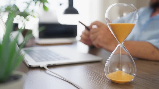 Focused hourglass with a blurred businessman working on a laptop, illustrating strategic thinking, planning, decision making and effective business management.