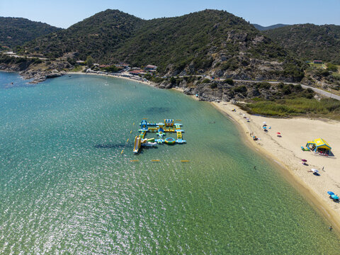 Aerial view of Sykias Beach with an inflatable water park floating in the clear turquoise sea, sandy coastline, and rocky hills in Paralia Sikias, Greece.