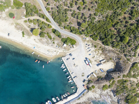 Aerial view of Sarti Beach featuring a small harbor with moored boats, a parking area, and surrounding Mediterranean vegetation, Greece.