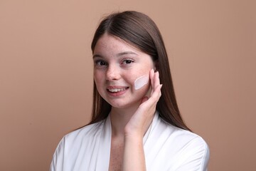 Obraz premium Beautiful girl with freckles applying cream on her face against dark beige background