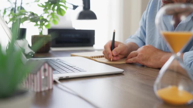 Businessman making notes while working at office desk with laptop and miniature house in background. Real estate planning, property investment, financial decisions and home ownership concept.