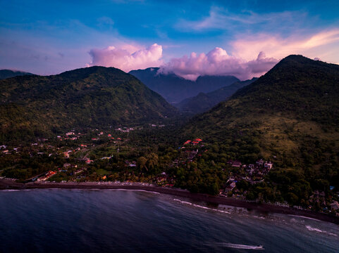 Aerial view of Amed in Abang, featuring lush green mountains, a black sand beach, and calm ocean waters under a vibrant sunset sky in Abang, Bali, Indonesia.