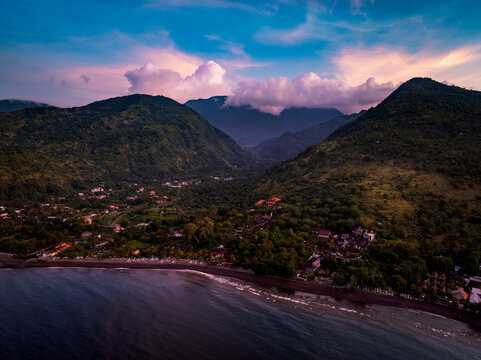 Aerial view of the coastal village of Amed, featuring dark volcanic sand beaches nestled against lush green mountains under a dramatic sunset sky in Abang, Bali, Indonesia.