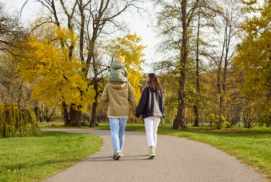 Happy family walking in autumn park together. Parents stroll a winding path as their daughter rides on dad's shoulders, passing yellow trees in soft sun. Simple moments strengthen family bonds.