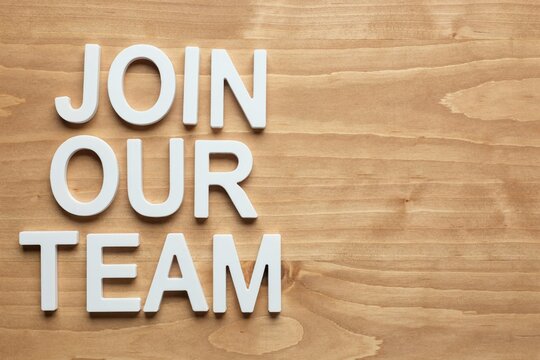 White 3 d letters spelling join our team on a light brown wooden surface