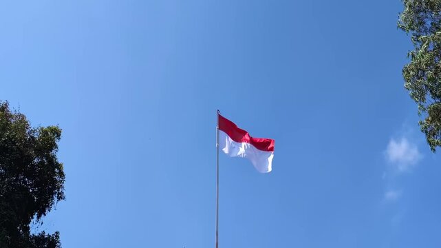 Indonesian national red and white flag waving on tall flagpole against clear blue sky, surrounded by festive independence day celebration decorations and lush green tree landscape.