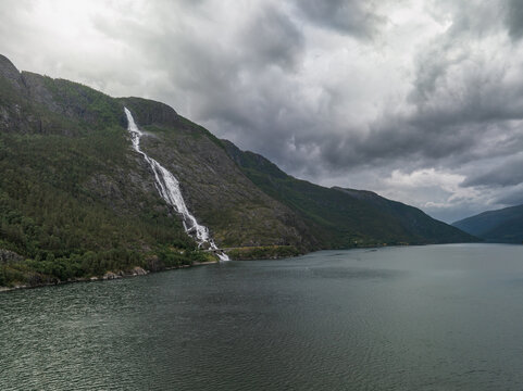 Aerial view of Langfoss waterfall cascading down the steep mountain slope into the deep waters of the fjord under a dramatic cloudy sky in Fjaera, Vestland, Norway.