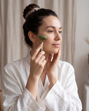 Woman in white robe using green jade facial roller on her cheek in soft natural light at home