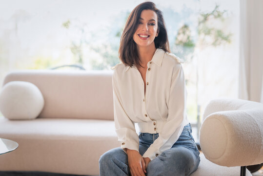 Full length of a cheerful smiling confident woman sitting on the couch in front of a floor-to-ceiling window