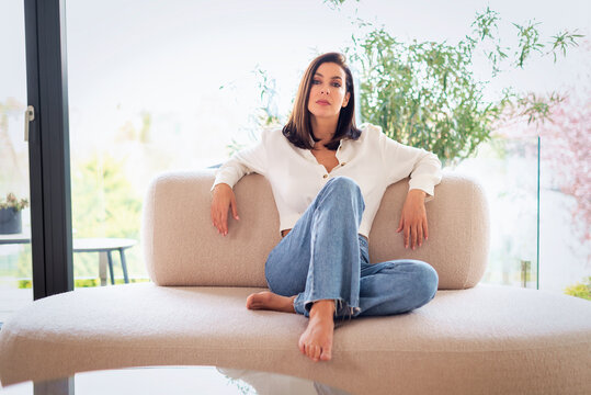 Full length of a cheerful smiling confident woman sitting on the couch in front of a floor-to-ceiling window