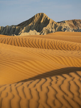 Aerial view of sand dunes with ripples and a rocky mountain in the background under a clear sky.