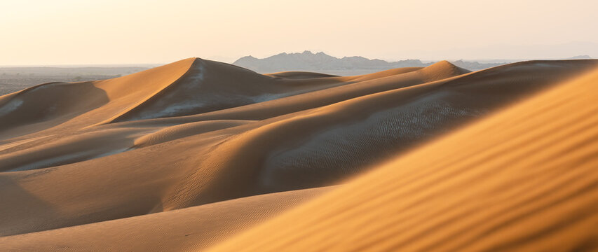 Aerial view of golden sand dunes with undulating ridges and soft shadows under a warm sunset sky.