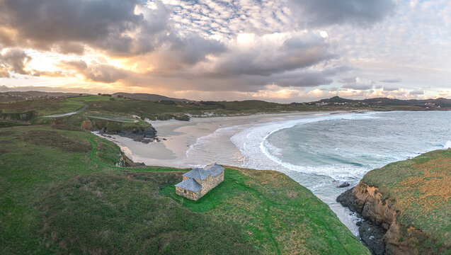 Aerial view of Ermita de Santa Comba on a green cliff overlooking the sandy beach and crashing waves under a cloudy sunset sky Covas, La Coruna, Spain.