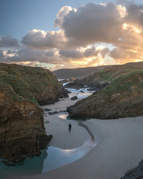 Aerial view of a lone person standing on a sandy beach near Ermita de Santa Comba nestled between rugged cliffs under a dramatic sunset sky Covas, La Coruna, Spain.
