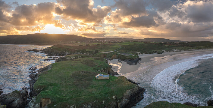 Aerial view of Ermita de Santa Comba on a green headland overlooking the sandy beach and crashing waves under a golden sunset sky Covas, La Coruna, Spain.