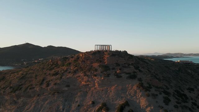 Greece - 01 June 2026: Aerial view of the ancient Temple of Poseidon at Cape Sounion with people walking around the ruins on a rocky cliff at sunset.