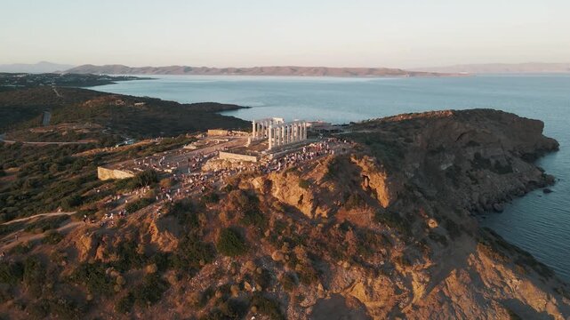 Sounion, Greece - 01 June 2026: Aerial view of the Temple of Poseidon at Cape Sounion with tourists gathered on the rocky cliffs at sunset.