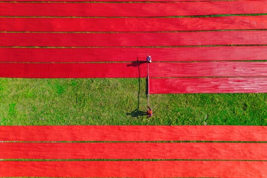 13 June 2026: Aerial view of workers laying out long strips of red fabric to dry on green grass, creating a vibrant pattern of parallel lines.