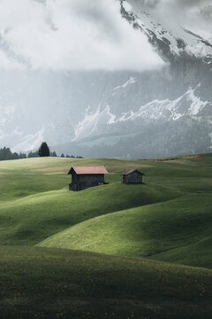 05 June 2024: View of two rustic wooden cabins on lush green rolling hills with a massive snow-capped mountain peak partially hidden by clouds.