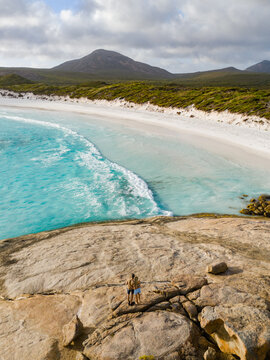 Aerial view of Hellfire Bay with turquoise water, white sand beach, and rocky foreground featuring a couple standing on the rocks in Cape Le Grand National Park, Western Australia, Australia.