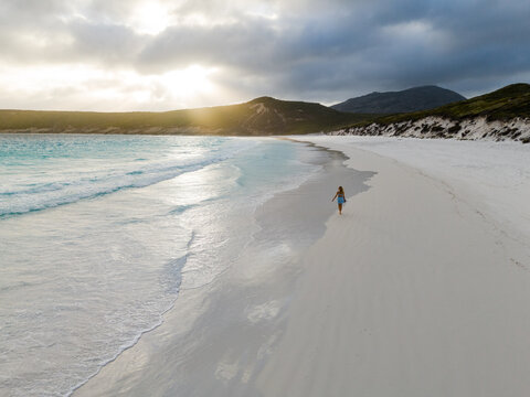 Aerial view of Hellfire Bay with a person walking along the white sandy beach and turquoise waves under a golden sunset in Cape Le Grand National Park, Western Australia, Australia.